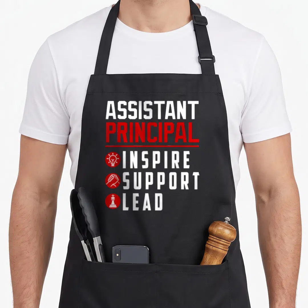 Inspire principal apron - Assistant Principal text with Inspire Support Lead icons and symbols in white and red — shown on a black cotton canvas apron against a white background