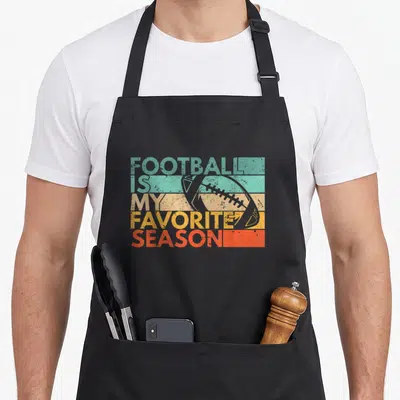 Funny season football apron - Stacked text FOOTBALL IS MY FAVORITE SEASON over colored stripes with grey football — shown on a black cotton canvas apron against a white background
