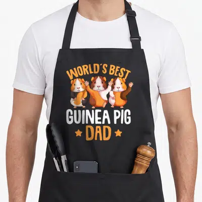 Pig guinea daddy apron - Three cartoon guinea pigs in orange and brown with text WORLD'S BEST GUINEA PIG DAD flanked — shown on a black cotton canvas apron against a white background