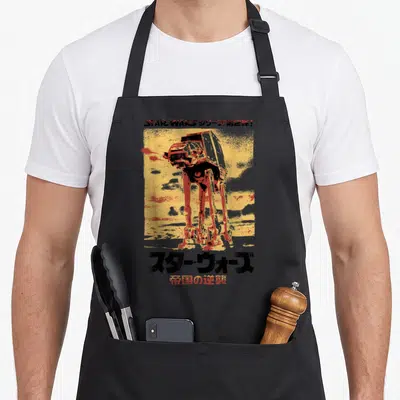 Empire japanese apron - AT-AT walker illustration on yellow background with Japanese Star Wars text — shown on a black cotton canvas apron against a white background