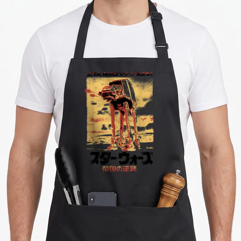 Empire japanese apron - AT-AT walker illustration on yellow background with Japanese Star Wars text — shown on a black cotton canvas apron against a white background