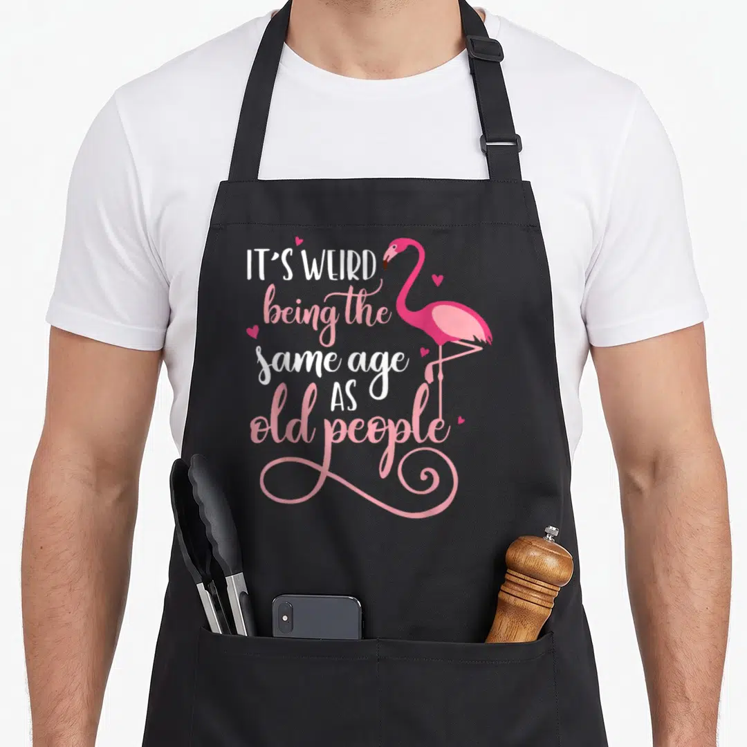 Age general weird aprons - Pink flamingo standing next to stacked white and pink text reading IT'S WEIRD being the same — shown on a black cotton canvas apron against a white background