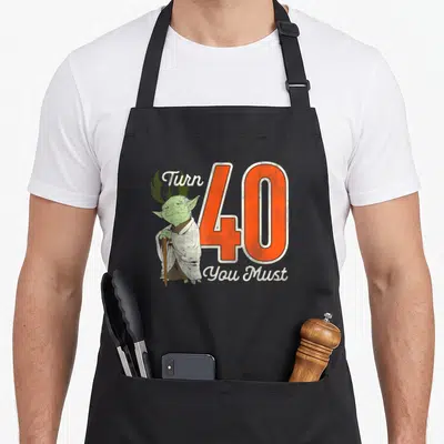 Yoda 40th apron - Green Yoda holding cane next to large orange 40 with Turn You Must text and green symbol — shown on a black cotton canvas apron against a white background