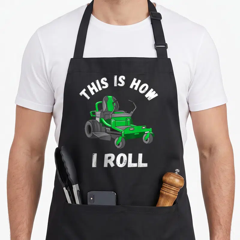 Mowing landscaper apron - Green riding lawn mower with arched white text THIS IS HOW I ROLL on grey background — shown on a black cotton canvas apron against a white background