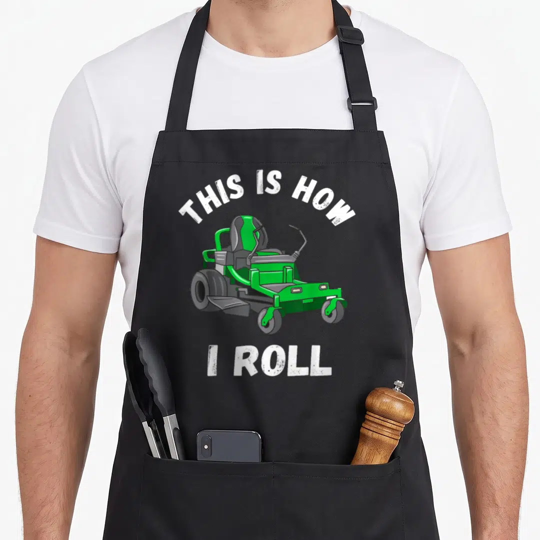 Mowing landscaper apron - Green riding lawn mower with arched white text THIS IS HOW I ROLL on grey background — shown on a black cotton canvas apron against a white background