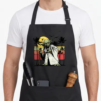 Yoda striped apron - Green character holding cane against yellow orange red striped sunset with black palm trees — shown on a black cotton canvas apron against a white background