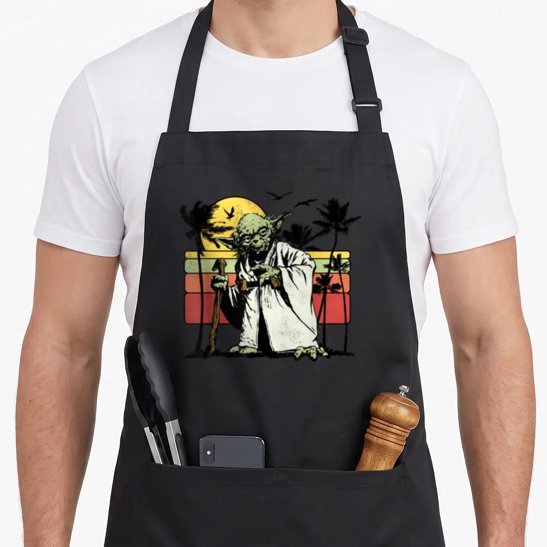 Yoda striped apron - Green character holding cane against yellow orange red striped sunset with black palm trees — shown on a black cotton canvas apron against a white background