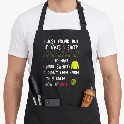 Joke knitting apron - Vertical text about sheep making wool sweaters with cartoon sheep and yarn ball — shown on a black cotton canvas apron against a white background