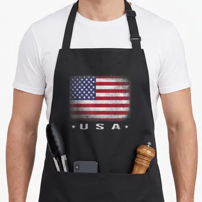 Faded 4th of july apron - Distressed American flag print above U S A text flanked by two white stars on red white — shown on a black cotton canvas apron against a white background