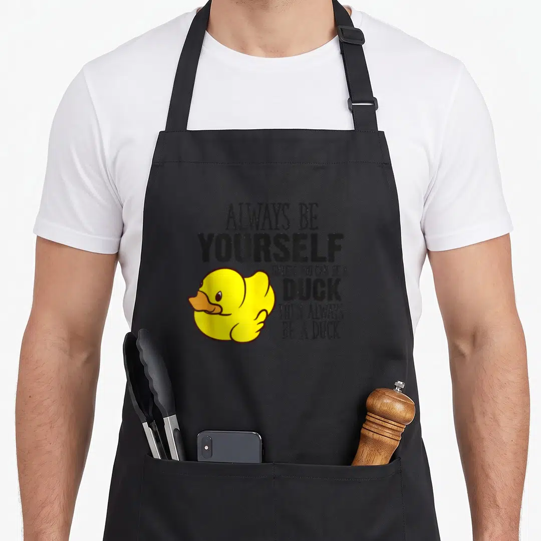 Alway yourself duck apron - Yellow cartoon duck next to stacked black text reading Always Be Yourself Unless You Can Be — shown on a black cotton canvas apron against a white background