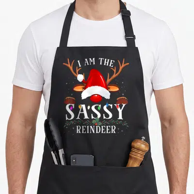 Sassy reindeer apron - Cartoon reindeer head with red hat between I AM THE SASSY REINDEER text with lights and holly — shown on a black cotton canvas apron against a white background