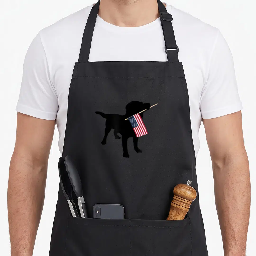 Holding 4th of july apron - Black dog silhouette holding red white and blue flag in mouth — shown on a black cotton canvas apron against a white background
