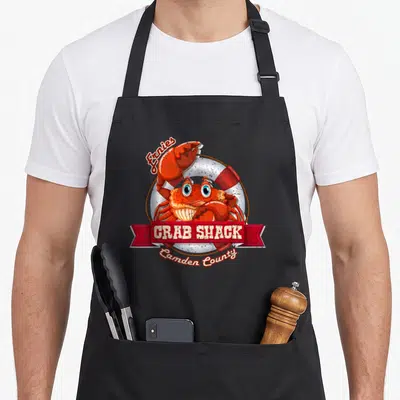 Shack crab apron - Red cartoon crab in front of lifebuoy with Ernies CRAB SHACK Camden County text — shown on a black cotton canvas apron against a white background