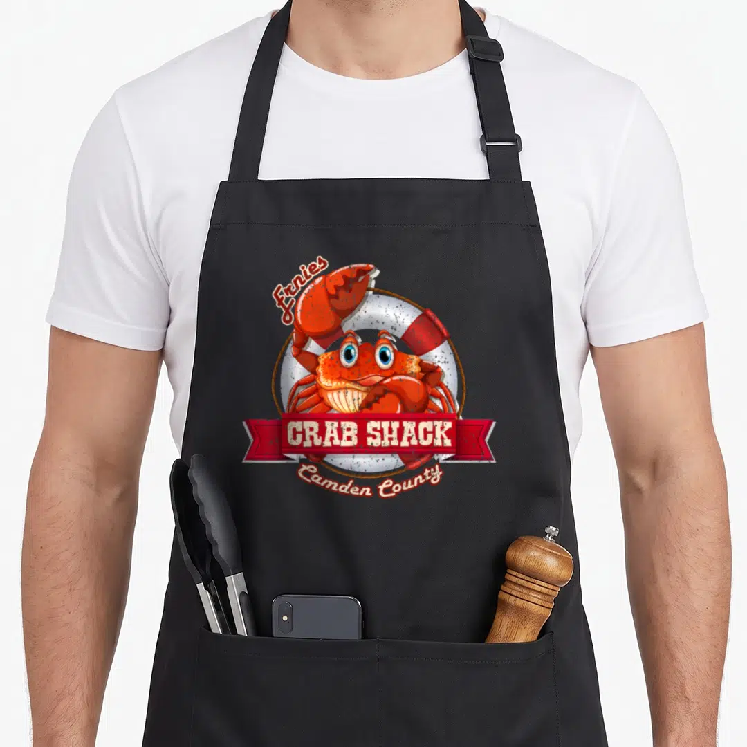 Shack crab apron - Red cartoon crab in front of lifebuoy with Ernies CRAB SHACK Camden County text — shown on a black cotton canvas apron against a white background