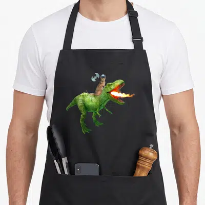 Galaxy cat apron - Brown cat in silver viking helmet riding green fire-breathing dinosaur holding axe — shown on a black cotton canvas apron against a white background