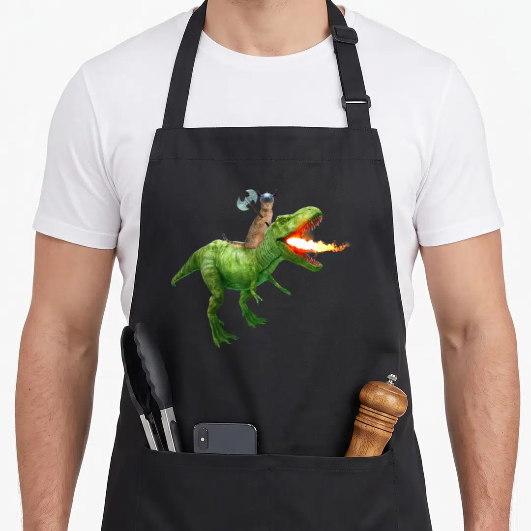 Galaxy cat apron - Brown cat in silver viking helmet riding green fire-breathing dinosaur holding axe — shown on a black cotton canvas apron against a white background