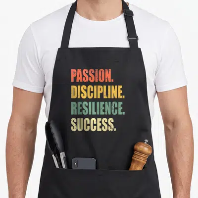 Passion influencer apron - Stacked distressed text reading Passion Discipline Resilience Success in orange yellow green — shown on a black cotton canvas apron against a white background