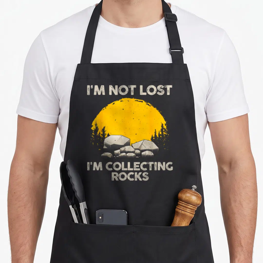 Rock collector apron - Distressed text I'm not lost I'm collecting rocks above yellow sun and grey rock cluster — shown on a black cotton canvas apron against a white background