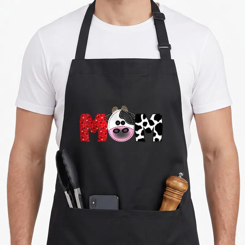 Daddy cow print apron - Cartoon cow head replaces O in MOM between red paisley M and spotted M — shown on a black cotton canvas apron against a white background