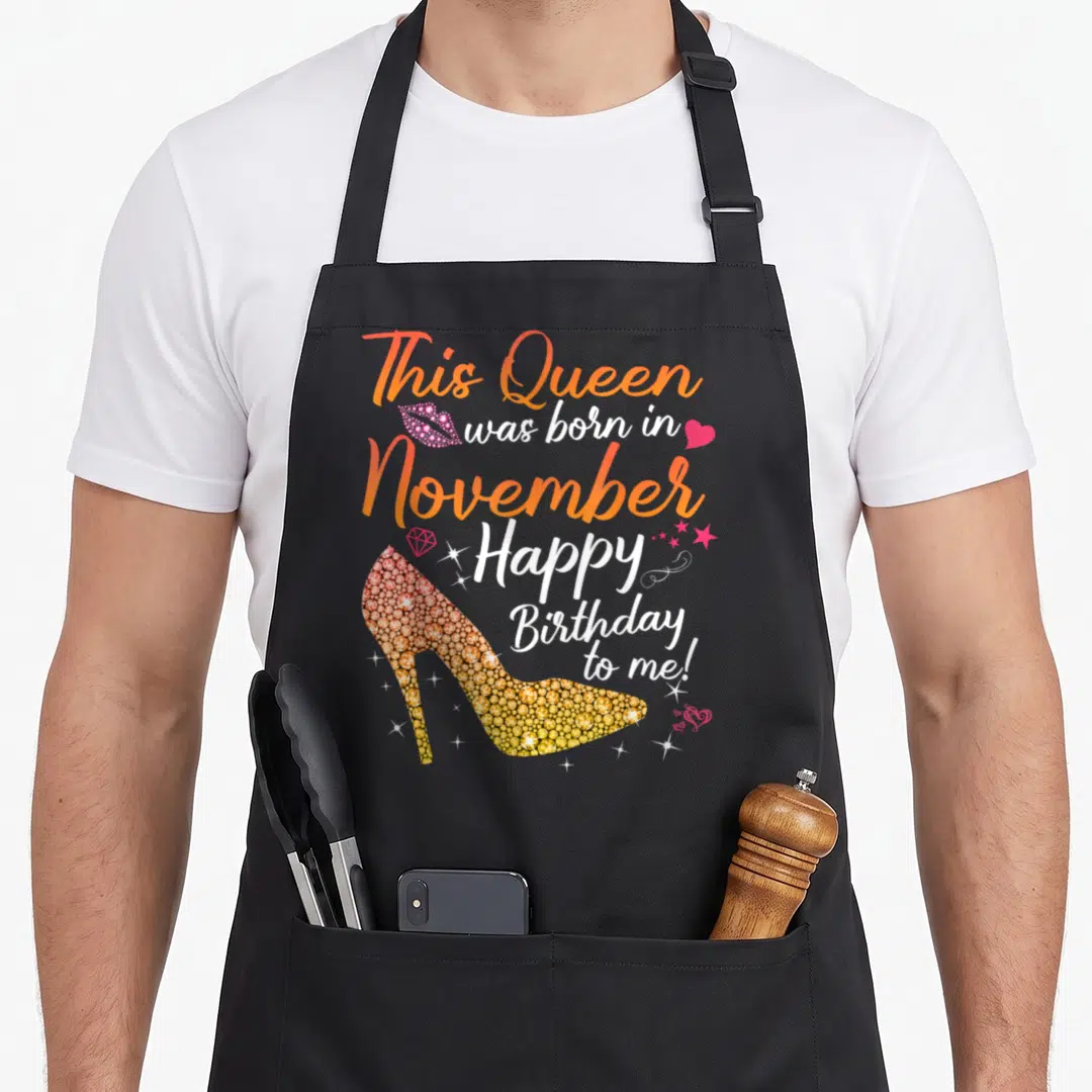 November baking queen apron - Glittery high heel shoe and stacked text reading This Queen was born in November Happy — shown on a black cotton canvas apron against a white background