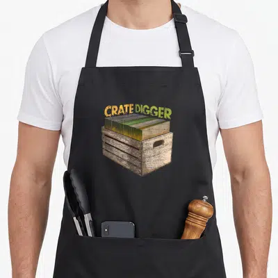 Geek vinyl apron - Arched CRATE DIGGER text above a distressed wooden crate filled with vinyl records in yellow — shown on a black cotton canvas apron against a white background