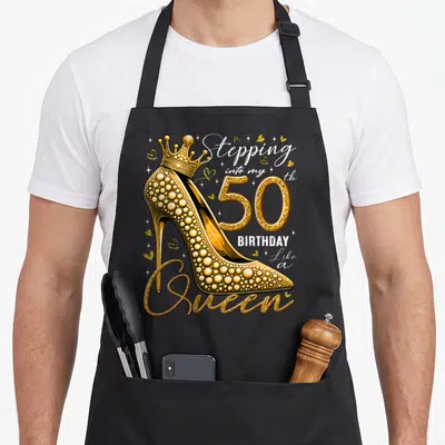 Heel stepping birthday apron - Gold glitter high heel with crown and 50th birthday quote on black apron — shown on a black cotton canvas apron against a white background