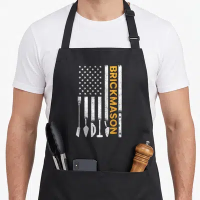 Brickmason american flag apron - Distressed white flag with masonry tool silhouettes and vertical orange BRICKMASON text — shown on a black cotton canvas apron against a white background