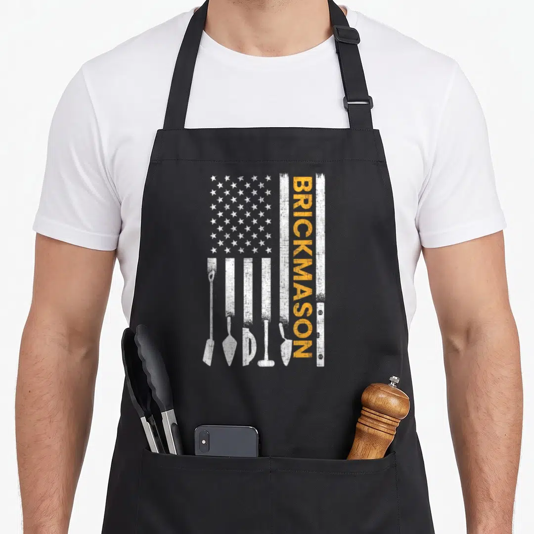 Brickmason american flag apron - Distressed white flag with masonry tool silhouettes and vertical orange BRICKMASON text — shown on a black cotton canvas apron against a white background