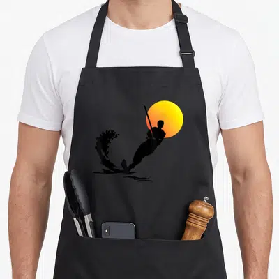 Water skiing apron - Black golfer silhouette swinging against yellow-orange sun with water splash — shown on a black cotton canvas apron against a white background