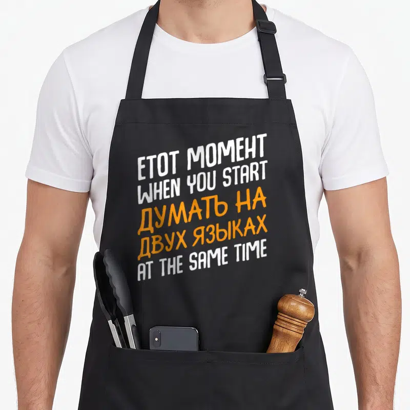 Start russian apron - Stacked white and orange bilingual text reading Etot Moment When You Start Think On Two Languages — shown on a black cotton canvas apron against a white background