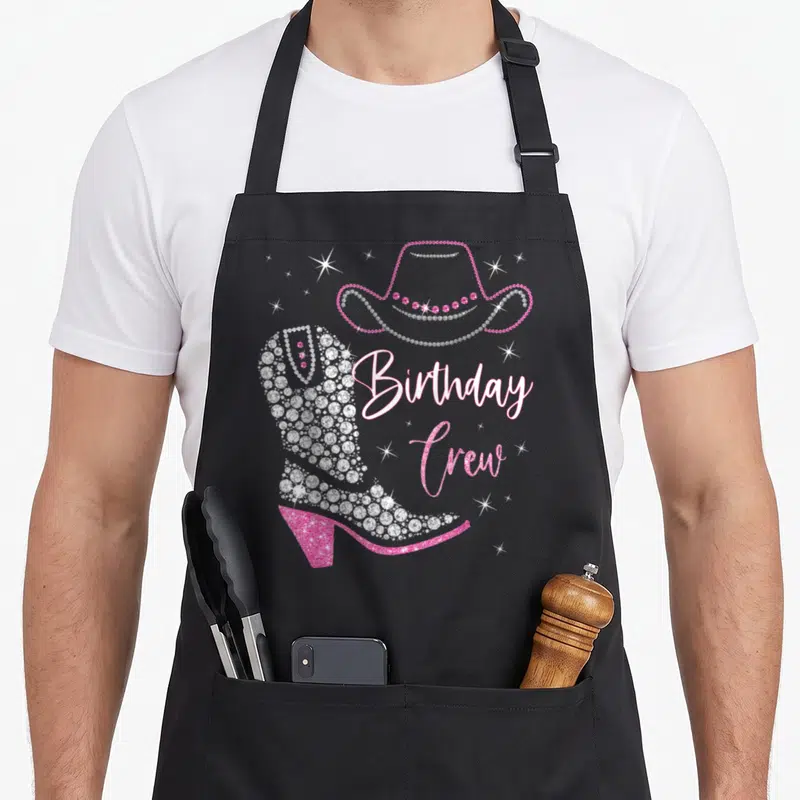 Crew cowgirl aprons - Rhinestone cowboy boot and hat with Birthday Crew text in pink and silver script surrounded — shown on a black cotton canvas apron against a white background