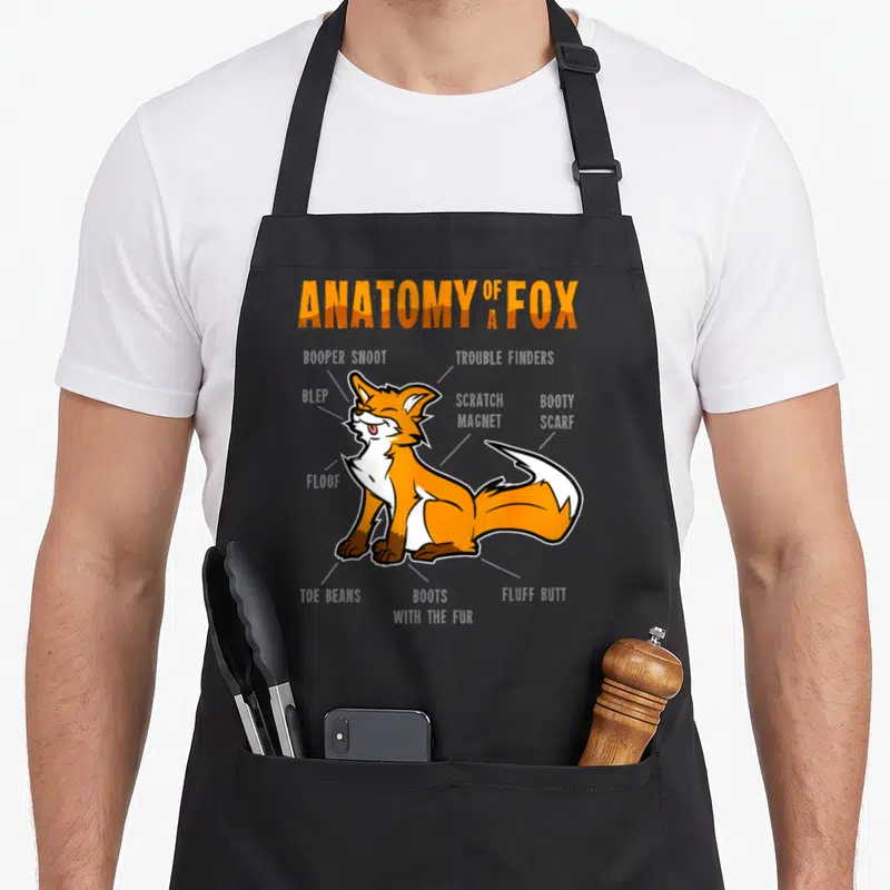 Fursona anatomy apron - Cartoon fox diagram labeled with terms like Blep, Toe Beans, and Floof in orange and grey — shown on a black cotton canvas apron against a white background