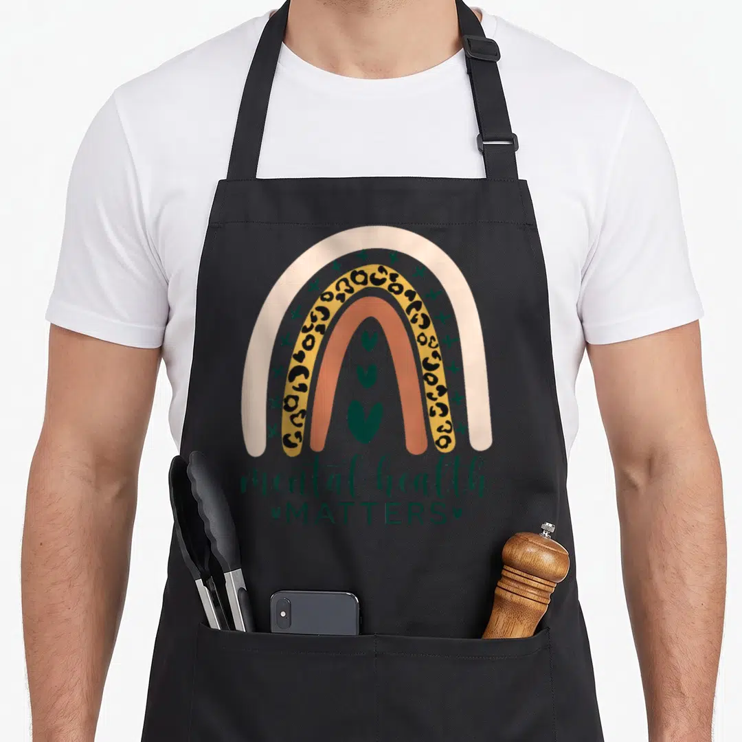 Mental leopard aprons - Rainbow with four arcs, three stacked hearts, mental health MATTERS text in beige green yellow — shown on a black cotton canvas apron against a white background
