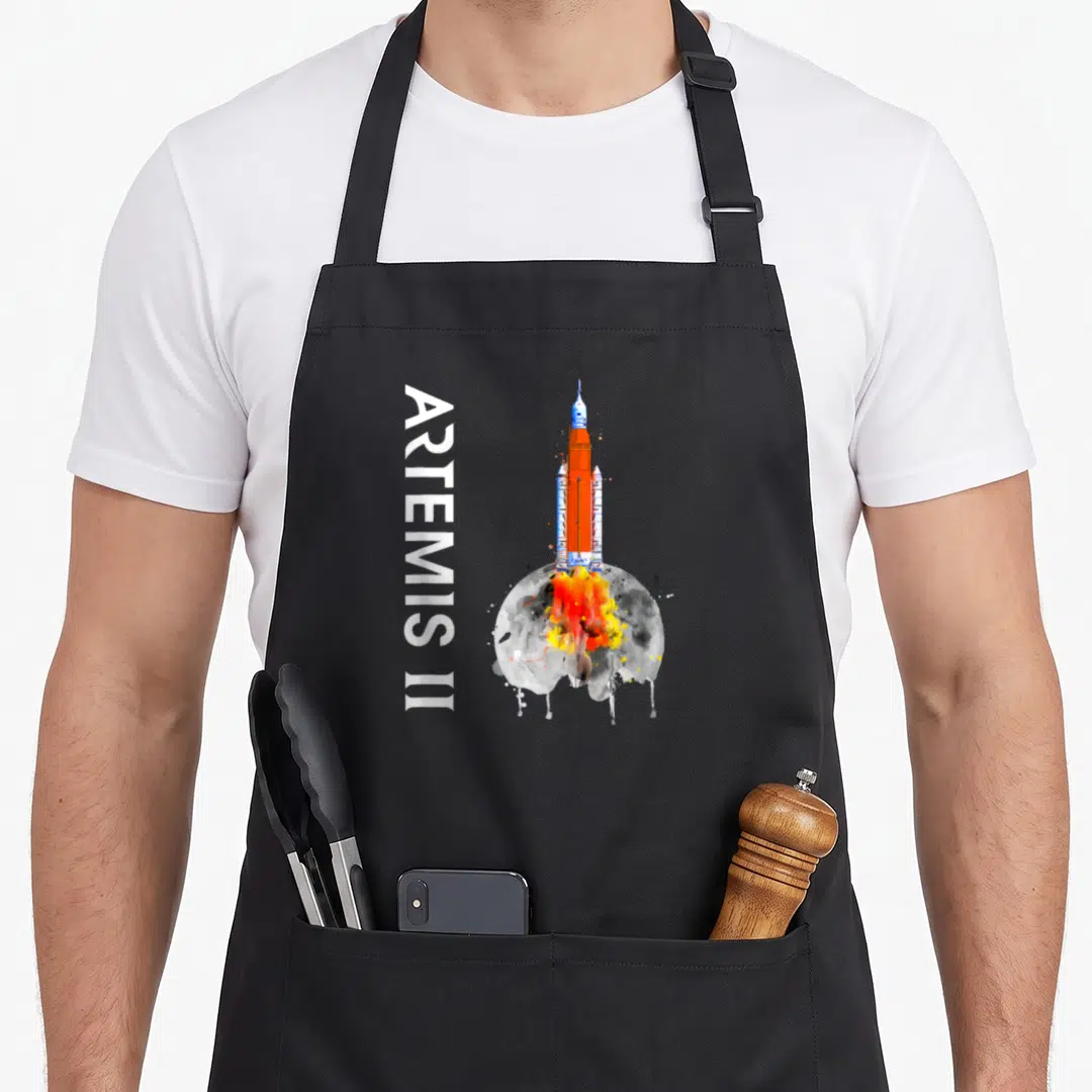 Rocket beyond apron - Vertical ARTEMIS II text beside orange rocket rising over grey moon with watercolor splashes — shown on a black cotton canvas apron against a white background