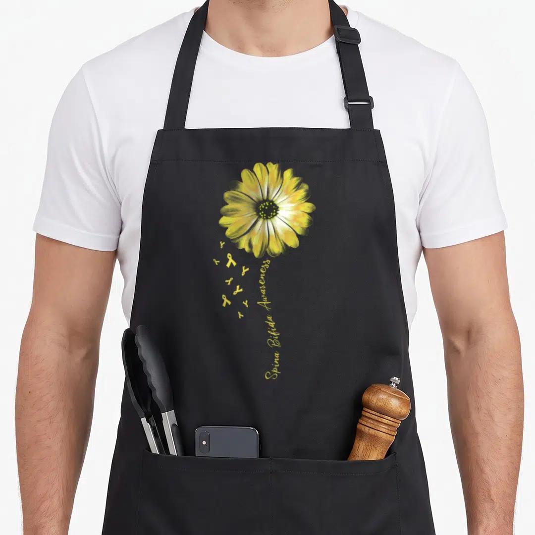 Spina daisy apron - Realistic yellow daisy with green accents and small ribbons forming a stem with text below — shown on a black cotton canvas apron against a white background