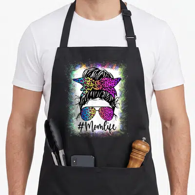 Rainbow leopard print cooking apron - Cartoon woman with bun, rainbow leopard bow, sunglasses, #Momlife text — shown on a black cotton canvas apron against a white background