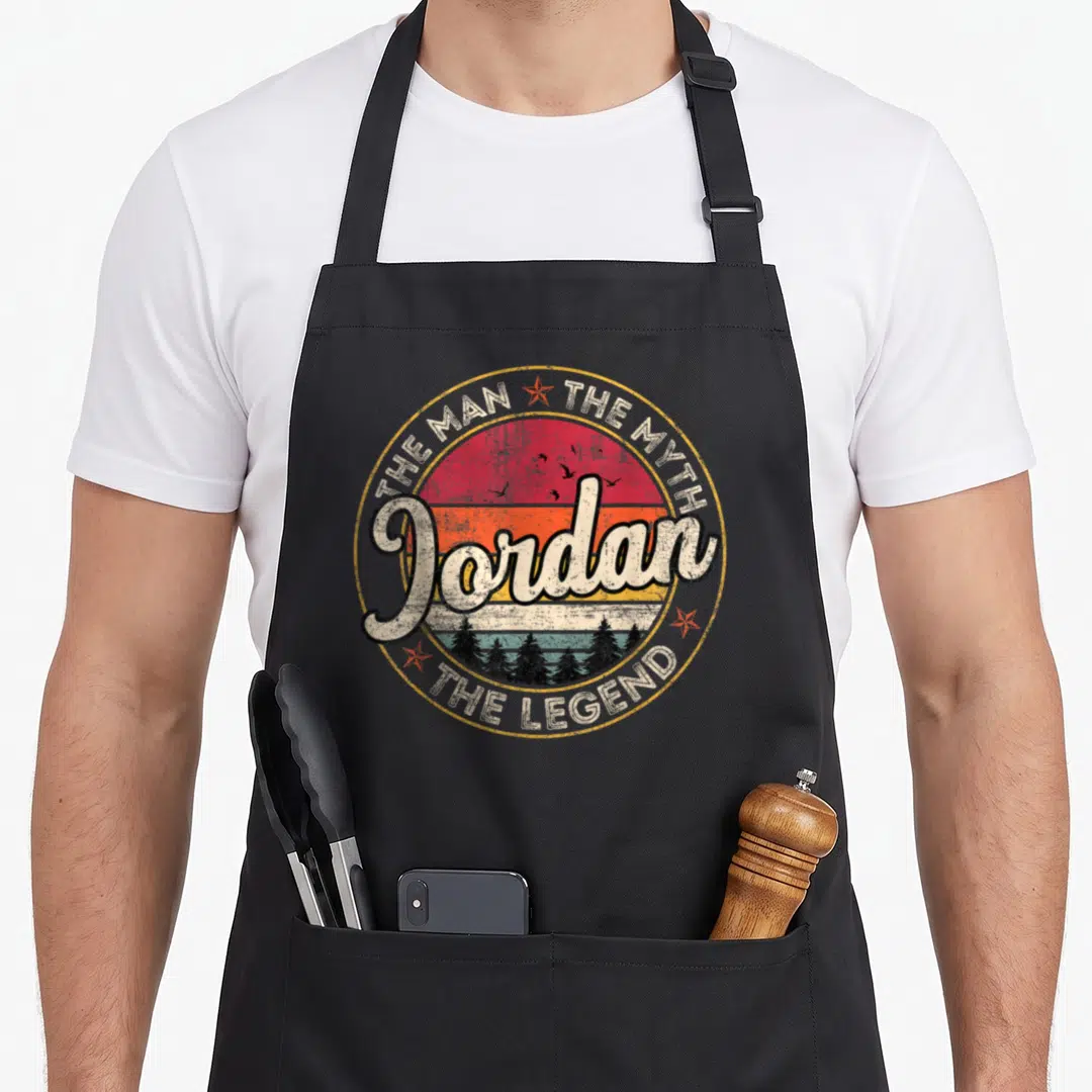 Myth special husband apron - Retro circular sunset design with Jordan script and birds flying over trees — shown on a black cotton canvas apron against a white background