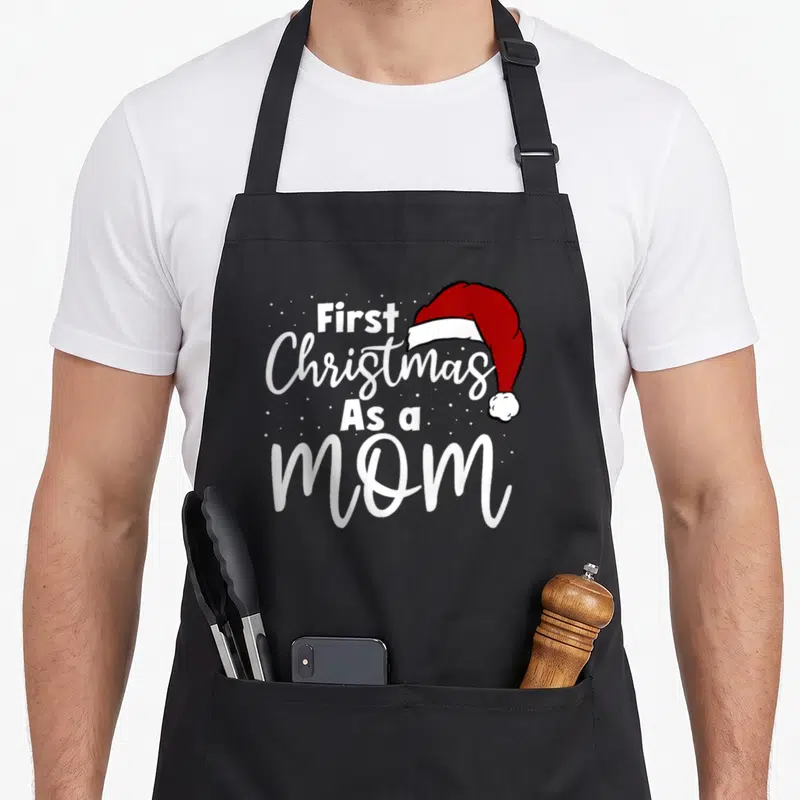 Family christmas aprons - White First Christmas As a mom text beside red Santa hat with snowflakes — shown on a black cotton canvas apron against a white background