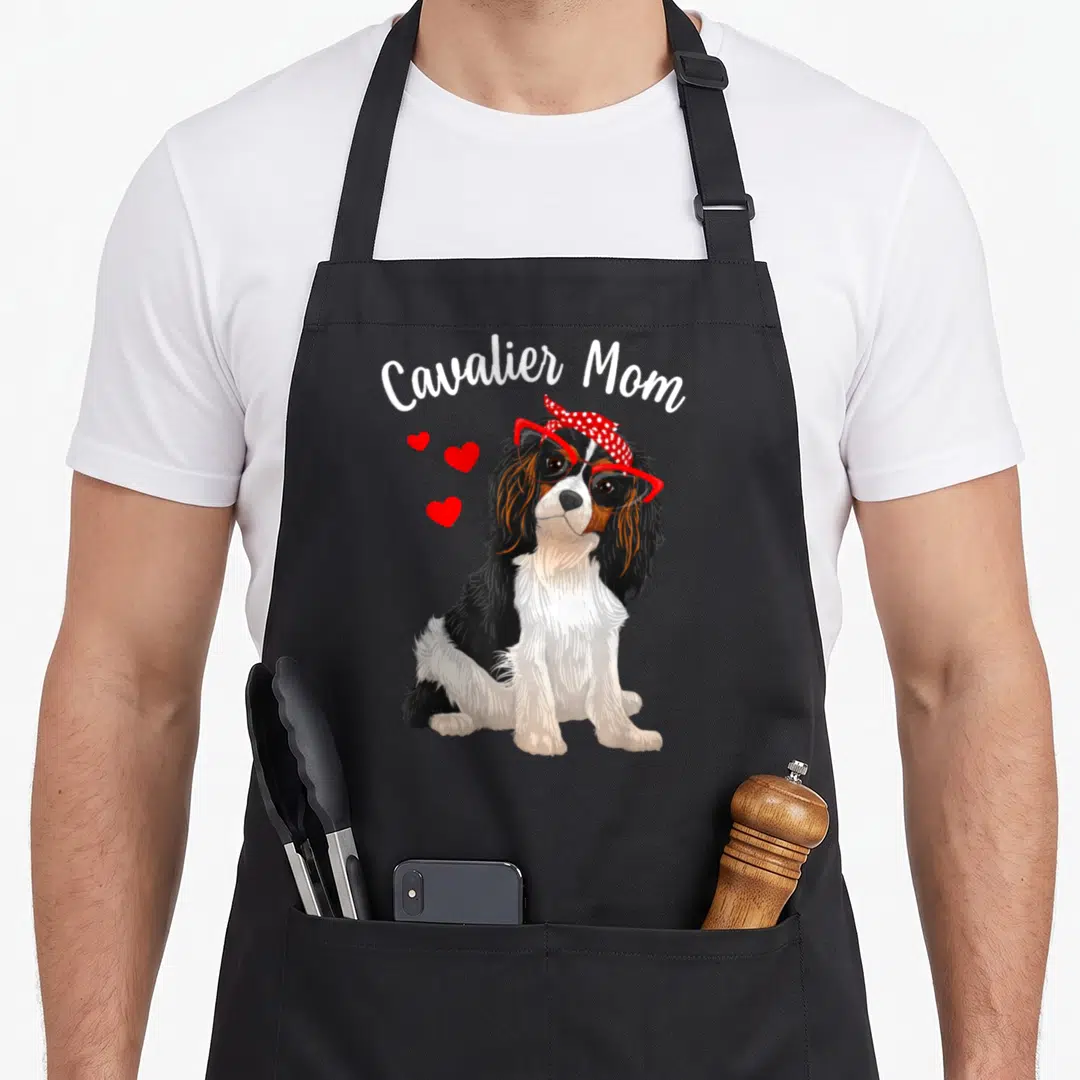 Cavalier charle spaniel king apron - Cavalier Mom text above a dog in red glasses and polka dot bandana with three — shown on a black cotton canvas apron against a white background