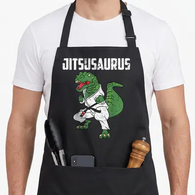 Jujitsu t-rex apron - Green dinosaur in white gi with black belt and JITSUSAURUS text — shown on a black cotton canvas apron against a white background