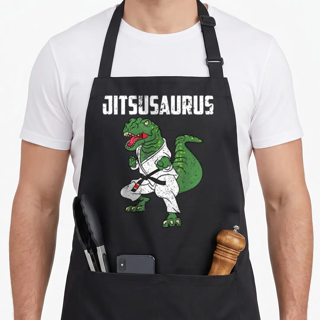 Jujitsu t-rex apron - Green dinosaur in white gi with black belt and JITSUSAURUS text — shown on a black cotton canvas apron against a white background
