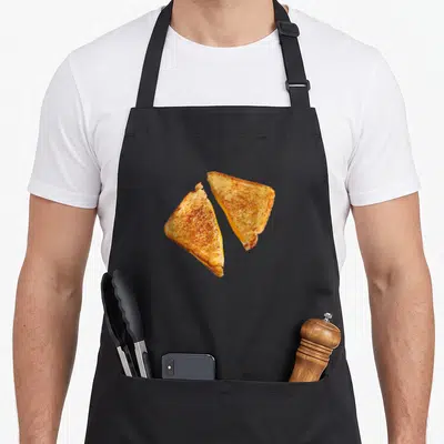 Grilled cheddar apron - Two diagonal halves of a golden brown grilled cheese sandwich with yellow cheese filling — shown on a black cotton canvas apron against a white background