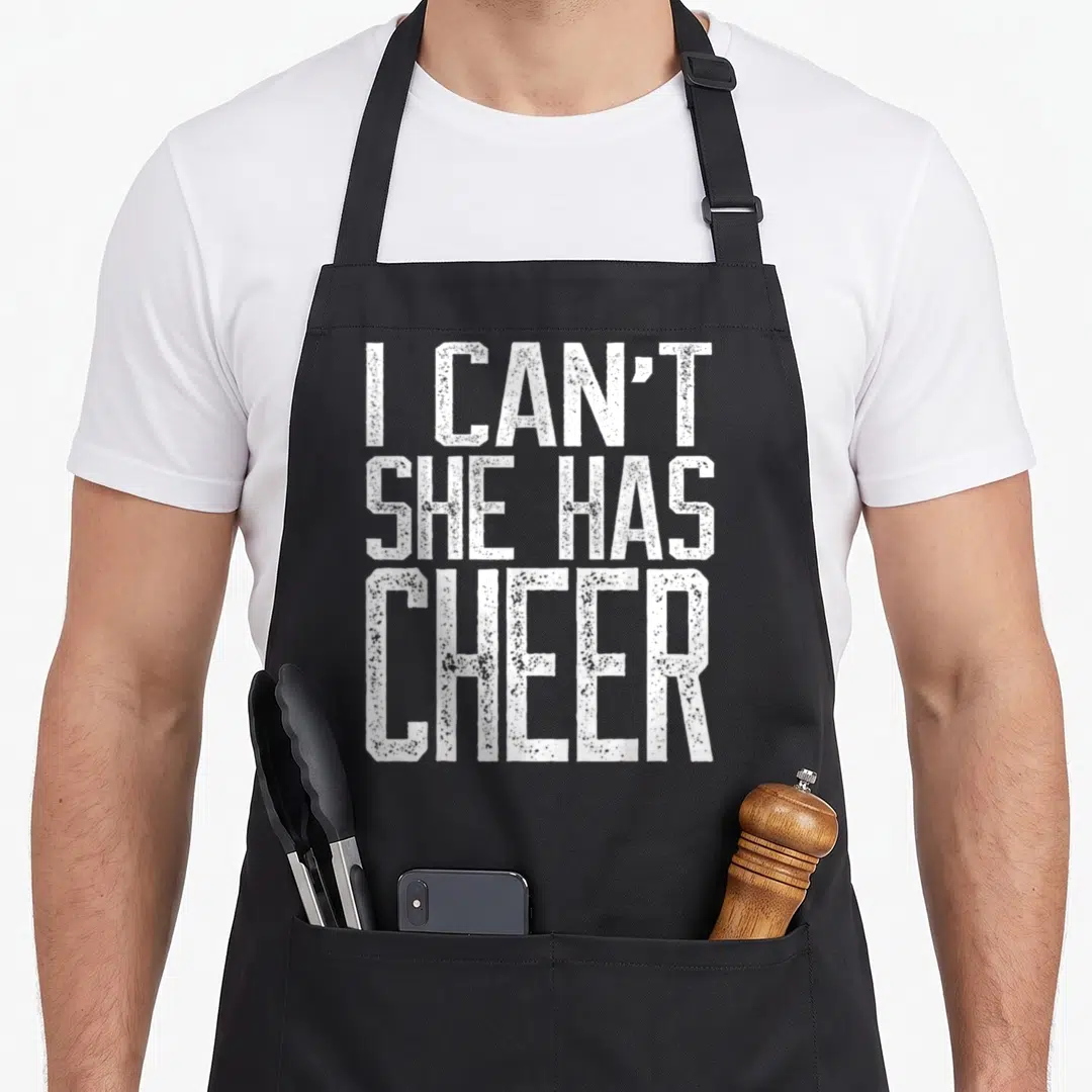 She cheerleading apron - White distressed text I CAN'T SHE HAS CHEER stacked vertically — shown on a black cotton canvas apron against a white background