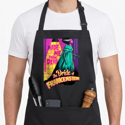 Dead bride apron - Bride of Frankenstein standing in spotlight with text Made Me From Dead in pink yellow teal purple — shown on a black cotton canvas apron against a white background