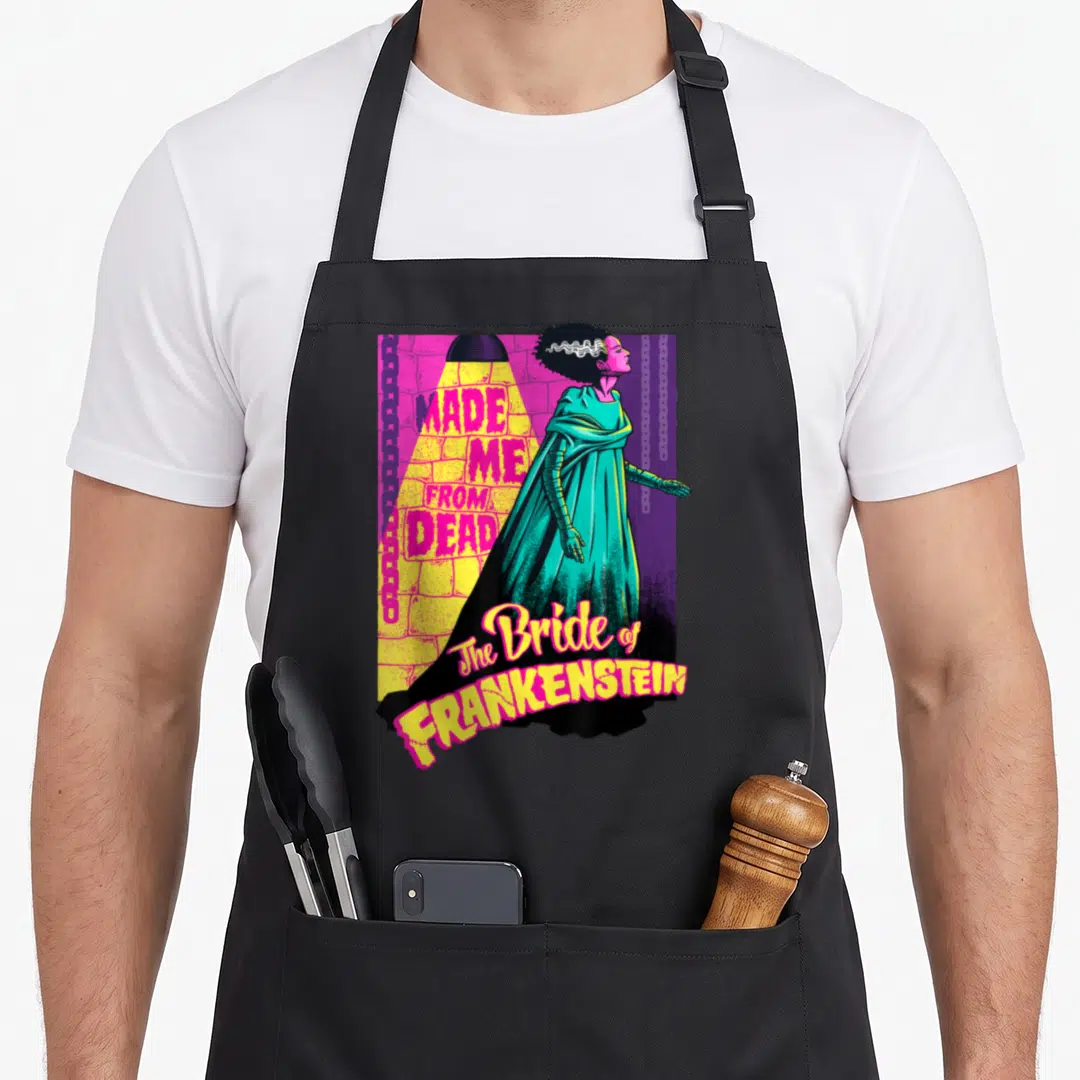 Dead bride apron - Bride of Frankenstein standing in spotlight with text Made Me From Dead in pink yellow teal purple — shown on a black cotton canvas apron against a white background