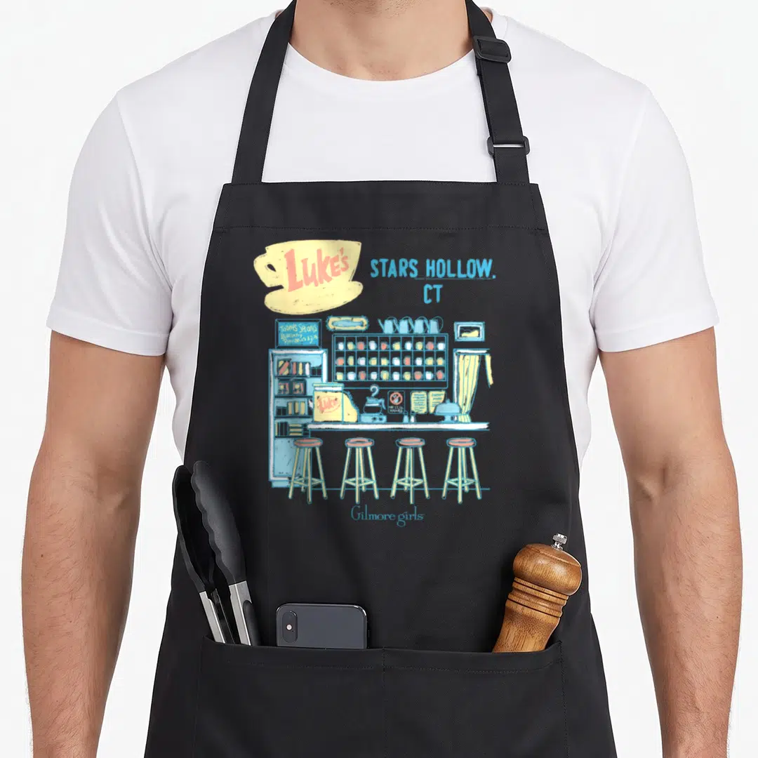 Gilmore pastel aprons - Cartoon sketch of Luke's diner counter with Stars Hollow text and pancake specials sign — shown on a black cotton canvas apron against a white background