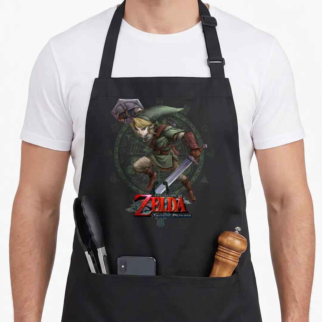 Twilight princess apron - Cartoon hero in green tunic with sword shield before grey circle, red Zelda logo text — shown on a black cotton canvas apron against a white background