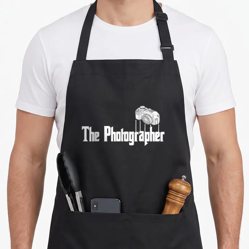 Vintage photography photographer apron - White line-art camera hanging above The Photographer text — shown on a black cotton canvas apron against a white background
