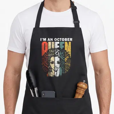 Queen birthday apron - I'M AN OCTOBER QUEEN text with woman head word cloud in red yellow teal white — shown on a black cotton canvas apron against a white background