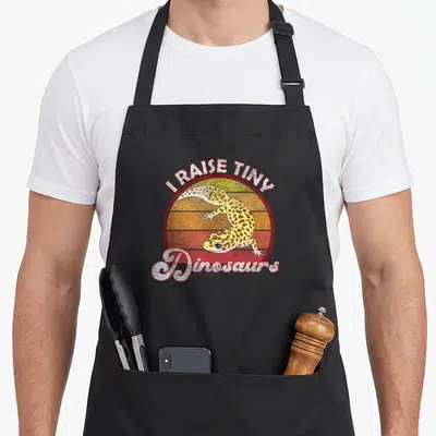 Dinosaur leopard apron - Retro sun graphic with leopard gecko and I RAISE TINY Dinosaurs text — shown on a black cotton canvas apron against a white background